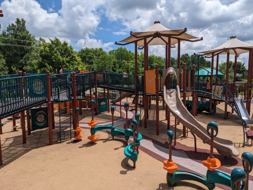 Colorful playground with slides, climbing structures, and shaded areas under a blue sky with fluffy clouds.