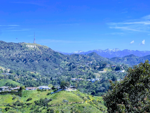 A panoramic view of the Hollywood sign on a hillside, surrounded by green hills and distant mountains under a clear blue sky.