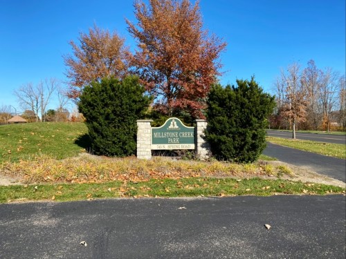 Sign for Millstone Creek Park, surrounded by trees and shrubs, with a clear blue sky in the background.