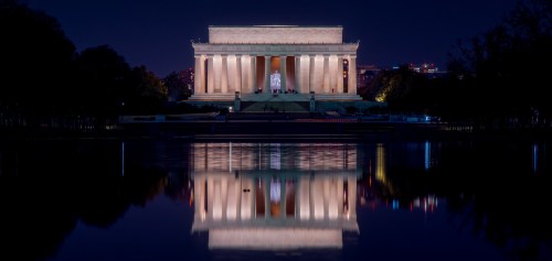 Lincoln Memorial illuminated at night, reflecting in the water below, surrounded by trees and city lights.