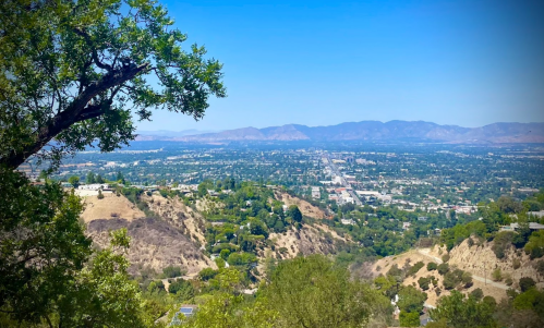 A panoramic view of a valley with hills, trees, and distant mountains under a clear blue sky.