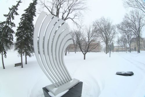 A modern sculpture stands in a snowy park, surrounded by bare trees and a winter landscape.