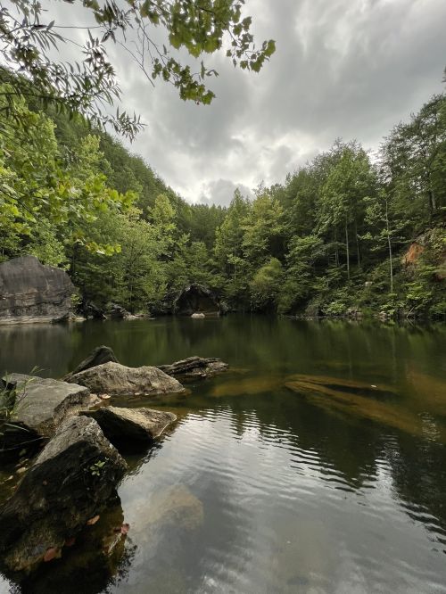 A serene lake surrounded by lush green trees under a cloudy sky, with rocks visible in the foreground.