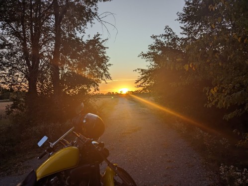 A motorcycle parked on a gravel path with a sunset in the background, surrounded by trees.
