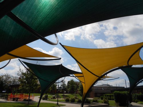 Colorful fabric canopies in green and yellow stretch across a park, with a blue sky and clouds in the background.