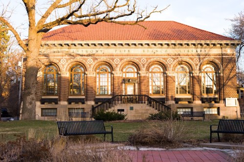 Historic library building with arched windows and a red tile roof, surrounded by benches and trees in a park setting.