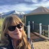 A woman with long hair and sunglasses smiles in front of a green yurt and mountains under a cloudy sky.