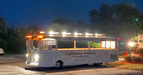 A brightly lit trolley bus parked at night, featuring the name "Lighthouse Trolleys" on its side.