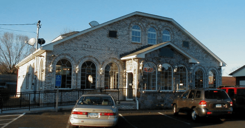 A brick building with large windows and a peaked roof, surrounded by parked cars in a parking lot.