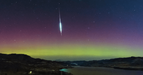 A vibrant night sky with a bright meteor streaking above a colorful aurora over a calm lake and hills.