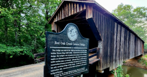 A wooden covered bridge with a historical sign in front, surrounded by trees and reflecting in the water below.
