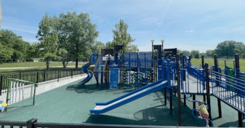 A colorful playground with slides and climbing structures, surrounded by green trees and a clear blue sky.
