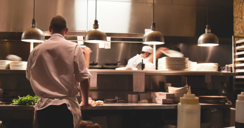 A chef in a busy kitchen prepares food, with other cooks working in the background and plates stacked nearby.