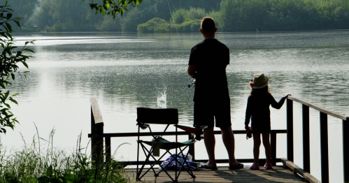 A person and a child stand on a dock by a calm lake, fishing and enjoying the serene nature around them.