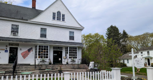 A white two-story building with an American flag, surrounded by greenery and a white picket fence.