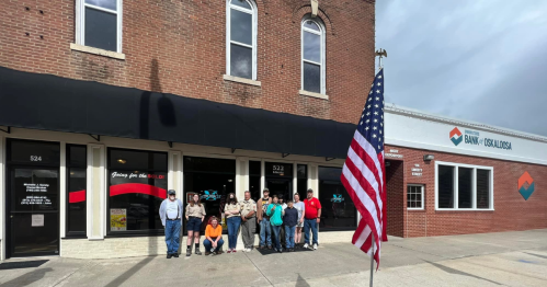 A group of people stands outside a brick building with an American flag in front, near a bank.
