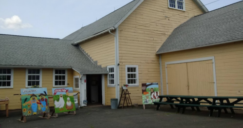 A yellow barn with an entrance, featuring colorful paintings on easels outside and picnic tables nearby.