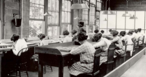 Black and white photo of women working at long tables in a well-lit room with large windows.