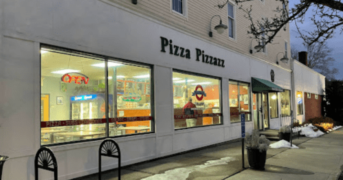 Exterior of "Pizza Pizzazz" restaurant at dusk, featuring large windows and a welcoming "OPEN" sign. Snow on the ground.