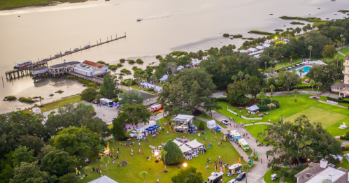 Aerial view of a waterfront festival with food stalls, people, and boats on the water, surrounded by greenery.