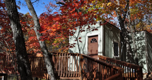 A small green cabin surrounded by vibrant autumn leaves and trees, with a wooden deck leading to the entrance.