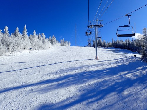 A snowy ski slope under a clear blue sky, with a ski lift and frosted trees in the background.