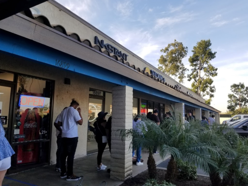 A line of people waiting outside a store called "Instant" under a blue sky, with palm trees nearby.