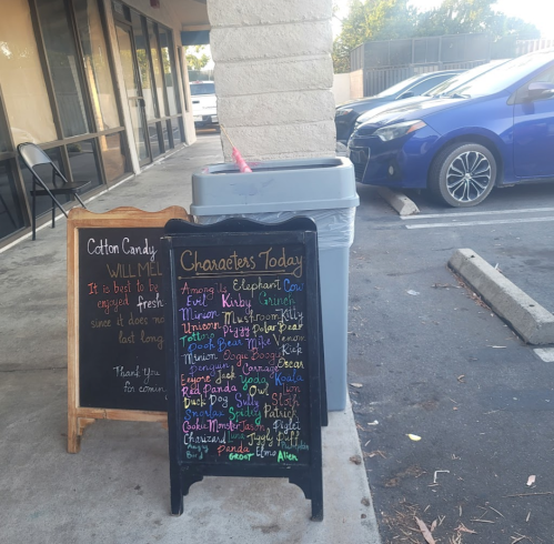 A sidewalk sign lists characters for the day, with a cotton candy stand nearby and a trash bin in the background.