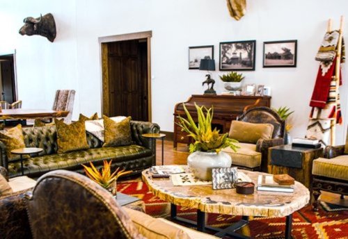 Cozy living room with a piano, rustic furniture, plants, and decorative art on the walls. Warm, inviting atmosphere.