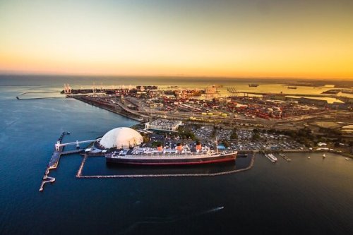 Aerial view of a harbor at sunset, featuring a large ship, industrial facilities, and calm waters.