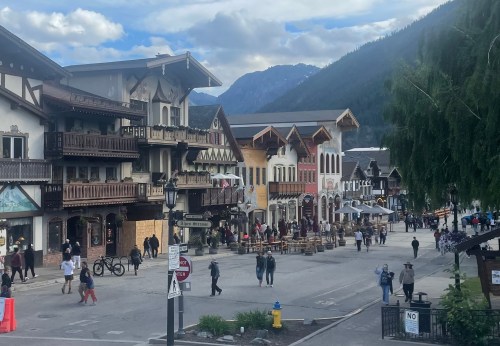 A charming street lined with alpine-style buildings, bustling with people and surrounded by mountains under a cloudy sky.