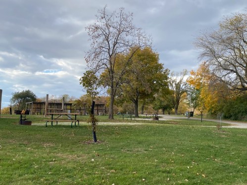 A park scene with trees, picnic tables, and a playground under a cloudy sky. Autumn foliage is visible.