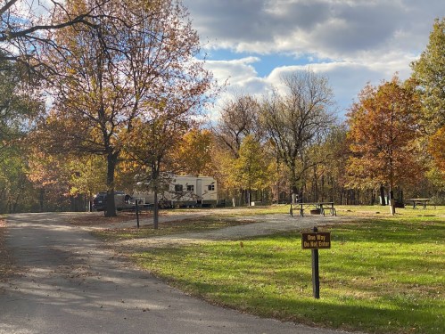 A scenic campground with autumn foliage, featuring an RV, picnic tables, and a "One Way Do Not Enter" sign.