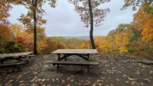 A wooden picnic table surrounded by colorful autumn foliage and a scenic view of a valley under a cloudy sky.