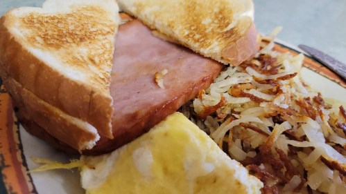 A plate with a toasted sandwich containing ham, alongside crispy hash browns and a slice of yellow egg.