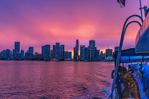 A vibrant sunset casts purple hues over a city skyline, viewed from a boat on the water.