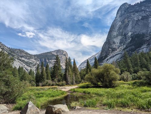 A scenic view of mountains and trees with a grassy area and a small pond under a partly cloudy sky.
