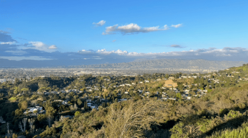 A panoramic view of a valley with hills, houses, and mountains under a clear blue sky with scattered clouds.