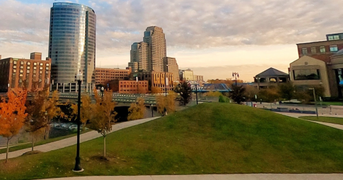 City skyline with modern buildings, a grassy hill, and autumn trees under a cloudy sky.