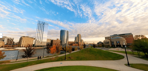 City skyline with modern buildings, a river, and colorful autumn trees under a blue sky with scattered clouds.