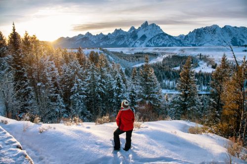 A person in a red jacket stands on a snowy hill, gazing at a mountain landscape during sunset.