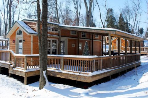 A cozy wooden cabin surrounded by snow, featuring a spacious deck and trees in a winter landscape.