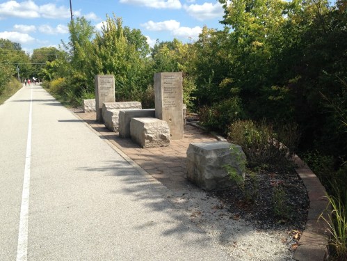 Stone benches and engraved markers line a paved path surrounded by greenery under a blue sky with clouds.