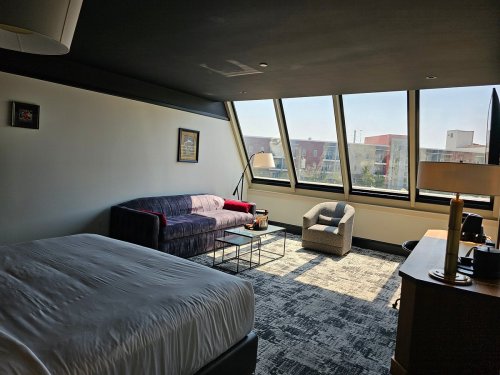 A modern hotel room featuring a bed, sofa, armchair, and large windows with a view of the outside.