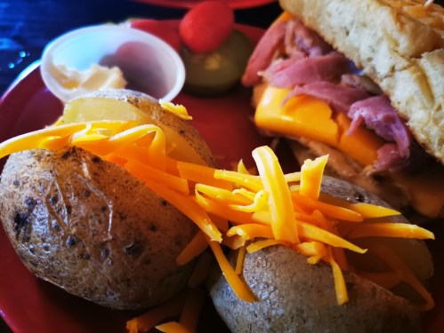 A plate with two baked potatoes topped with shredded cheese, a sandwich, and condiments in small cups.