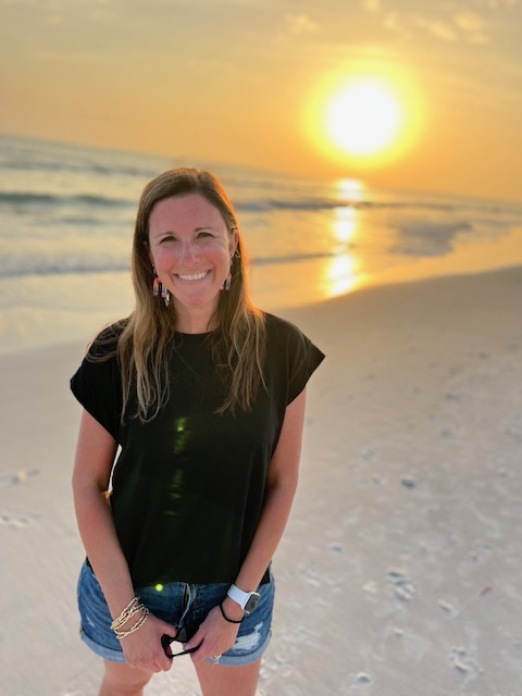 A woman stands on a beach at sunset, smiling with the sun setting over the ocean in the background.