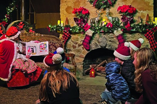 A woman in a festive outfit reads a story to children by a decorated fireplace with holiday decorations.