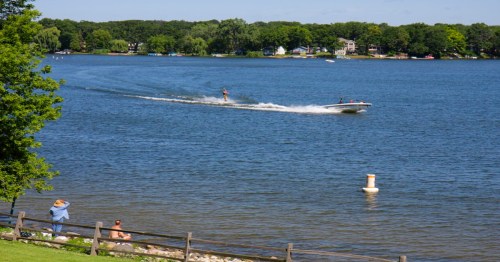 A person water skiing on a lake, with trees and houses in the background and people relaxing by the shore.