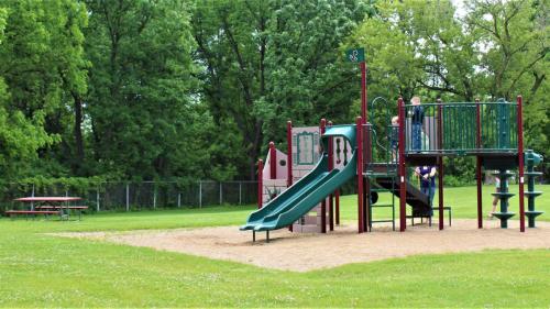 A playground with a slide and climbing structure, surrounded by green grass and trees, with picnic tables nearby.