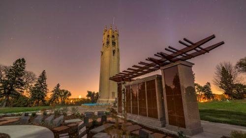 A tall tower stands against a twilight sky, surrounded by trees and a landscaped area with seating.